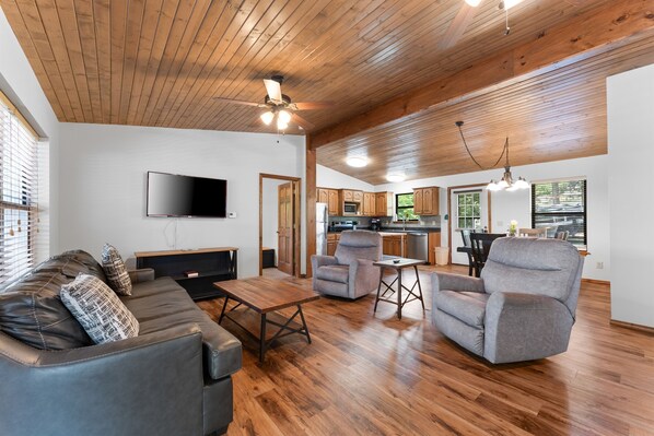A spacious living room with a wooden ceiling, a flat-screen TV mounted on the wall, a leather sofa, and a gray armchair.