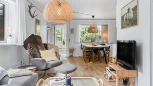 A cozy living room with a grey sofa, a wooden coffee table, and a television. There is a dining area with a table and chairs, and a large window providing a view of the outdoors. - 2 bedroom stunning home in Væggerløse (Væggerløse)