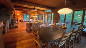 A log cabin interior with a large dining table, wooden chairs, a fireplace, and large windows offering a view of the outdoors. - The Cabin on Fontana Lake
Lake Front Paradise in the Smokies (Robbinsville)