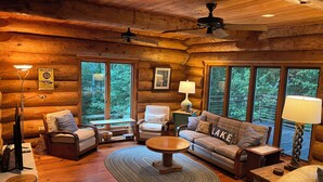A cozy log cabin interior with a fireplace, wooden furniture, and large windows offering a view of the forest. - The Cabin on Fontana Lake
Lake Front Paradise in the Smokies (Robbinsville)