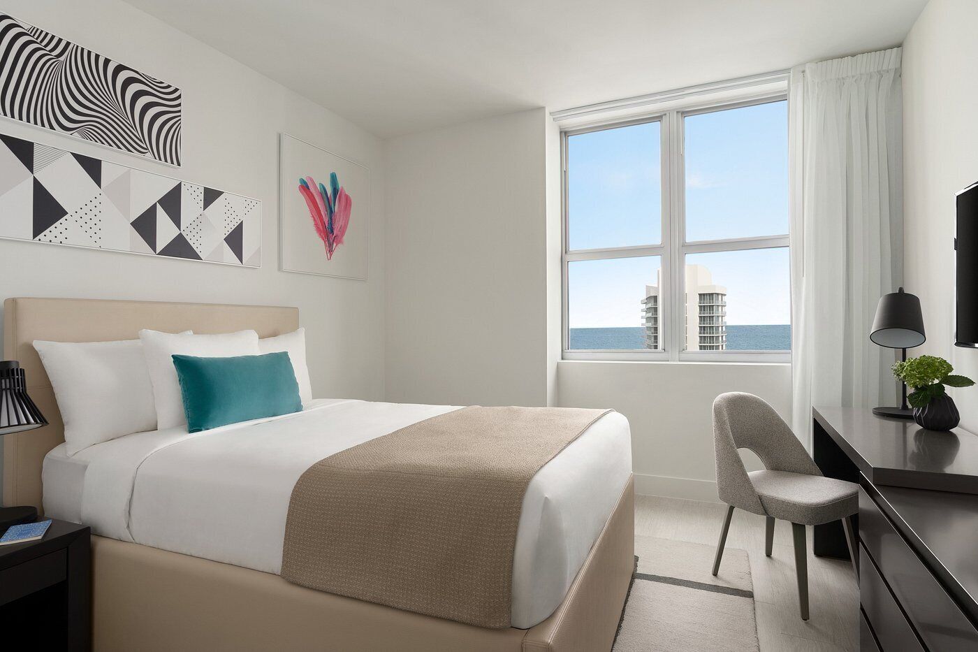 A hotel room with a bed, a desk, a chair, and a large window with a view of buildings and the sea.
