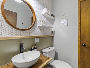 Un baño con un tocador de madera, un lavamanos blanco, un inodoro, un espejo con marco de madera y un estante con productos de aseo.