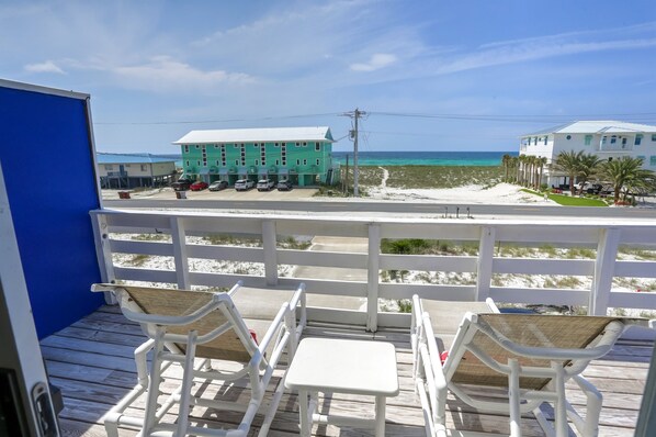 Eine Holzterrasse mit weißen Stühlen und einem Tisch mit Blick auf einen Strand und ein türkisblaues Meer, im Hintergrund sind Gebäude zu sehen.