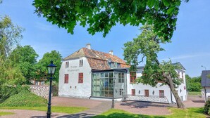 Un bâtiment blanc avec des encadrements de fenêtres rouges et un toit recouvert de tuiles, entouré de verdure et bordé d’un chemin de gravier.