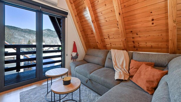 A cozy room with a wooden ceiling, a grey sofa, a small round table, and a large window offering a view of a snowy landscape. - Beautiful home in Jasenak with WiFi (Jasenak)