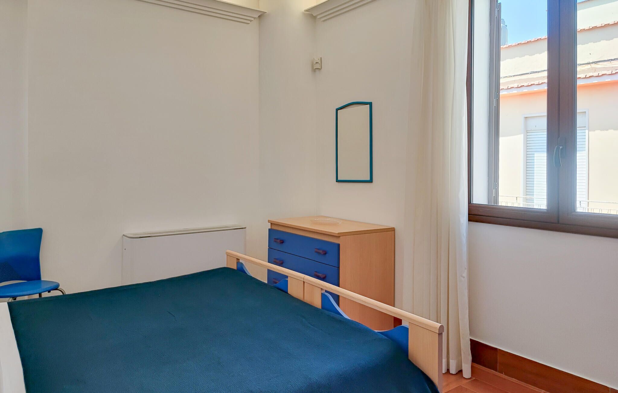 A bedroom with a bed, a wooden dresser, a blue chair, a window with curtains, and a wall-mounted mirror.