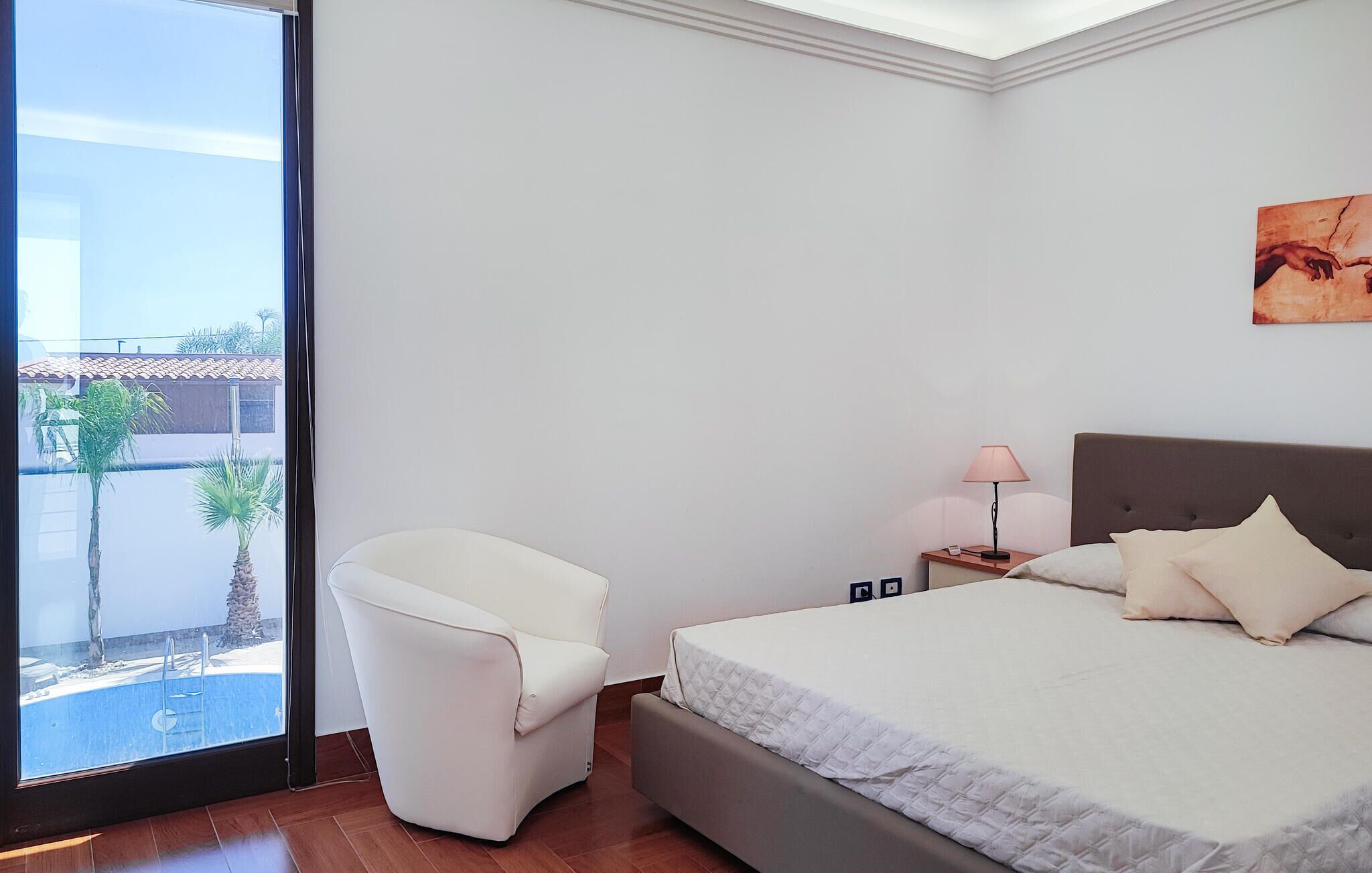 Een slaapkamer met een bed, een stoel, een nachtkastje en uitzicht op een buitenzwembad.