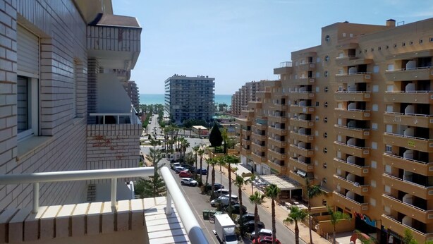 A high-rise building with multiple balconies, a street with parked cars, and palm trees.