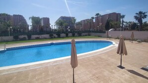 A swimming pool with a curved edge, surrounded by a tiled deck. There are two umbrellas and a fence enclosing the area.