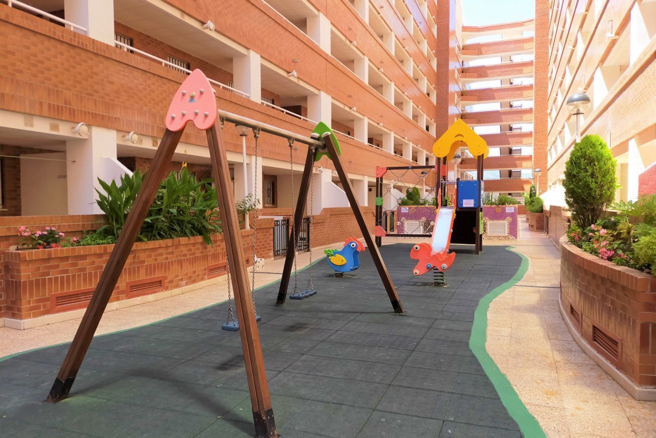 Ein Spielplatz mit Schaukeln und Rutschen, umgeben von einem mehrstöckigen Backsteinhaus.
