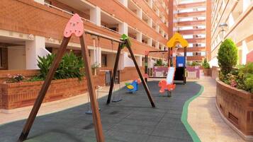 Ein Spielplatz mit Schaukeln und Rutschen, umgeben von einem mehrstöckigen Backsteinhaus.