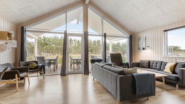 A spacious living area with a large sliding glass door leading to an outdoor patio. There are multiple seating options including a sofa, chairs, and a dining table. The room features wooden flooring and a high ceiling with a sloped design.
