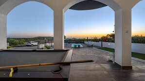 Mesa de bilhar em área externa coberta, com vista para uma piscina e um pôr do sol.