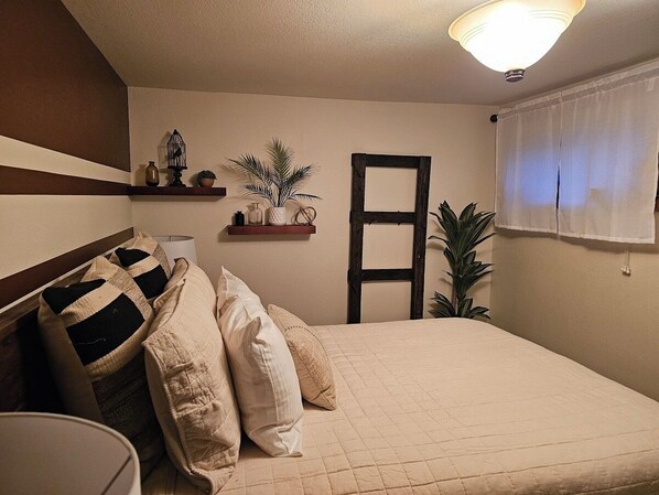 Un dormitorio con cama, almohadas, una mesita de noche, lámpara, estante con adornos, ventana con cortinas y una lámpara en el techo.