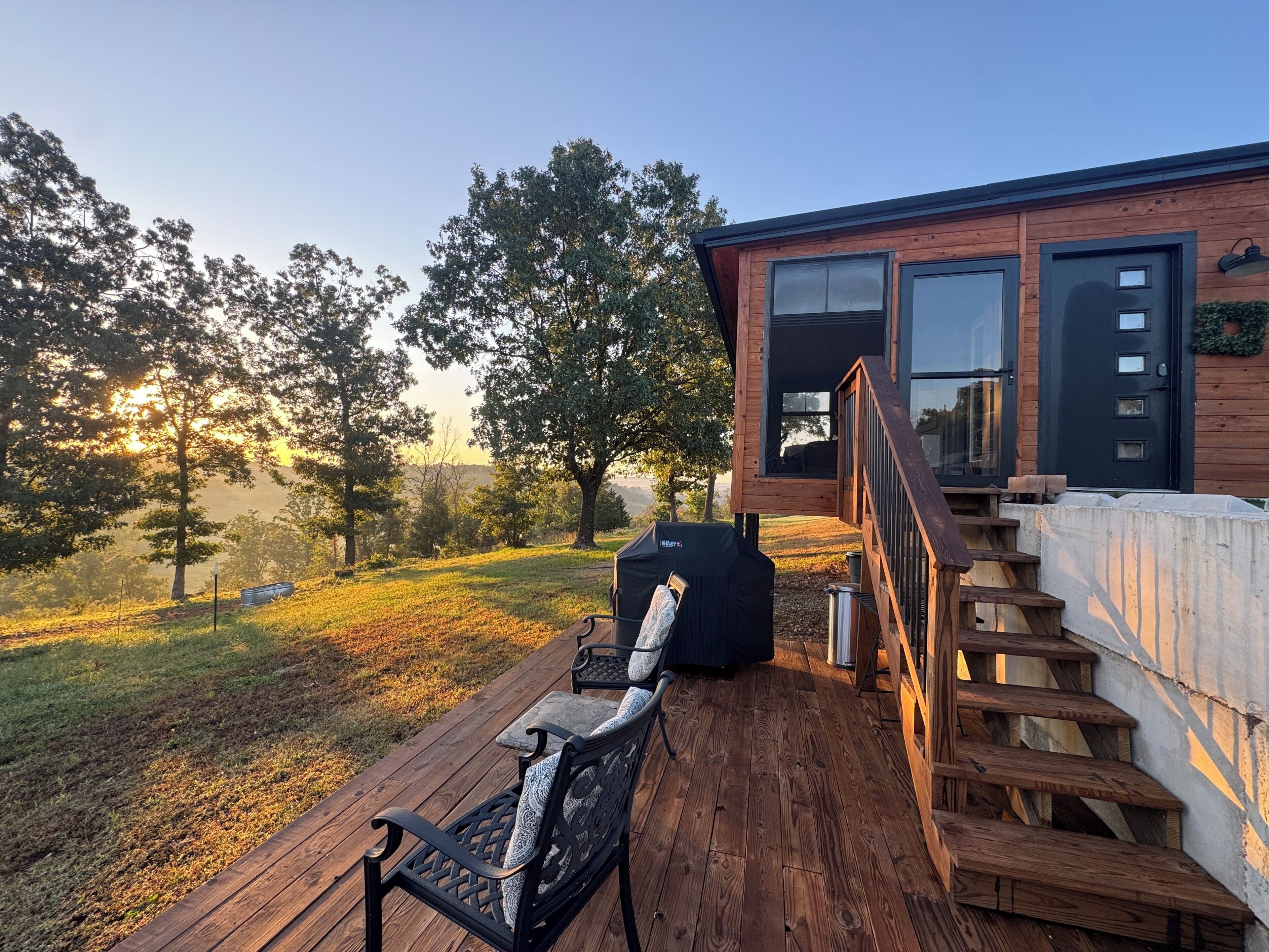 Una terraza de madera con mobiliario exterior, una escalera que conduce a un edificio con ventanas amplias y vistas a árboles y colinas al fondo.