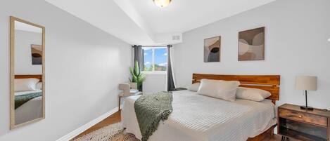 A modern bedroom with a large bed, a bedside table, a nightstand, a mirror, and a window with a view.