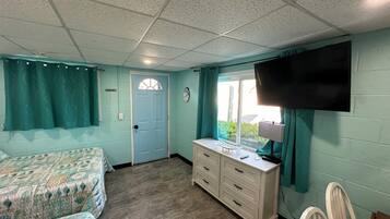 A room with teal walls, a white ceiling, a bed with patterned bedding, a dresser, a TV mounted on the wall, and a window with curtains.