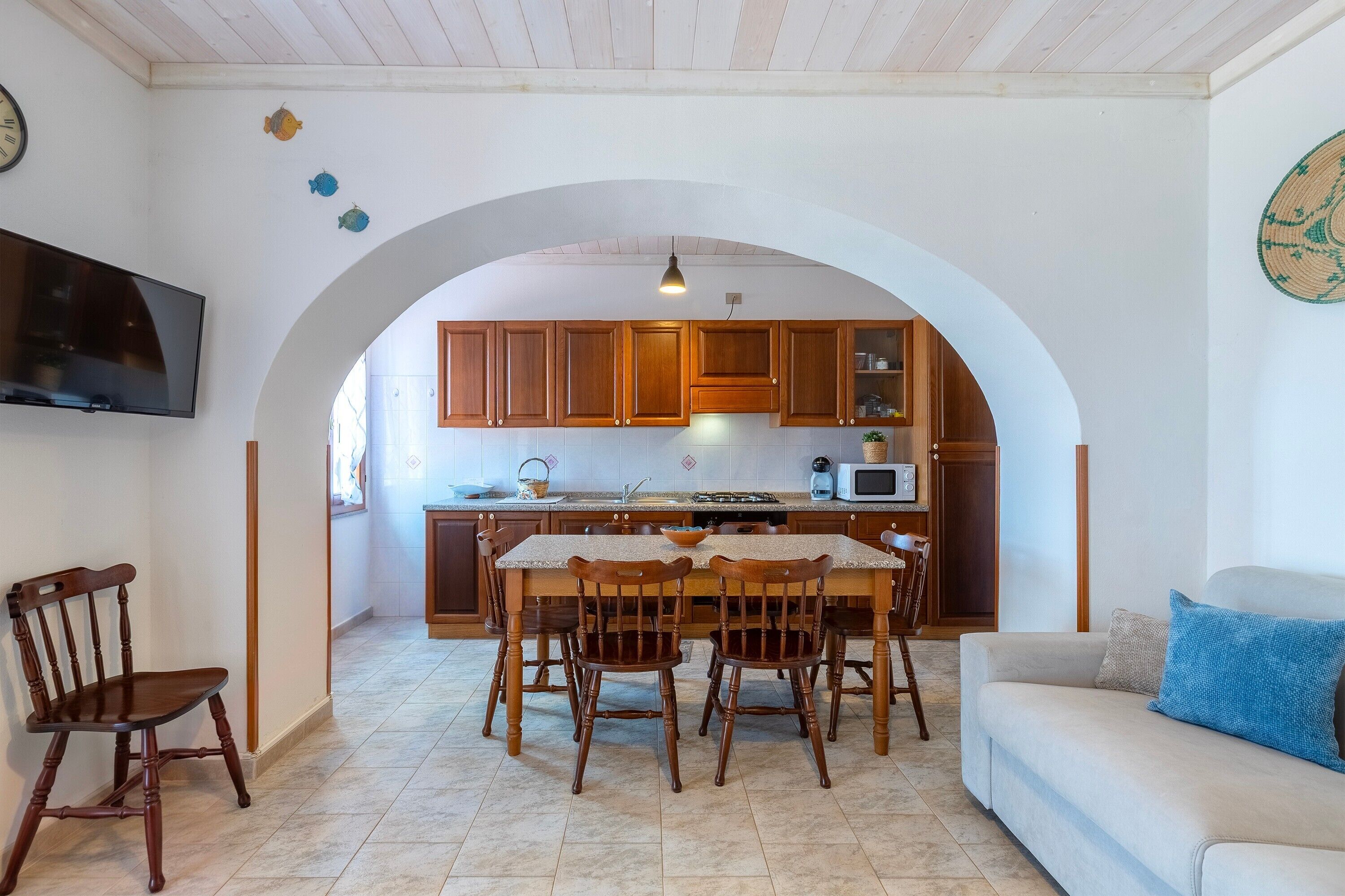 Une cuisine avec des meubles en bois, une table à manger avec des chaises, un téléviseur fixé au mur et une porte en arc donnant sur une autre pièce.