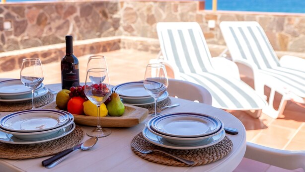 A table set for a meal with plates, cutlery, and a bottle of wine, overlooking a stone wall and the sea.
