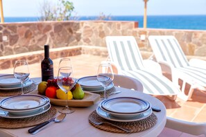 A table set for a meal with plates, cutlery, and a bottle of wine, overlooking a stone wall and the sea.