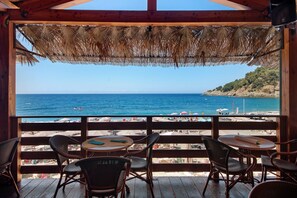 Terraza de madera con sillas de mimbre y una mesa, con vistas a una playa con sombrillas y gente.
