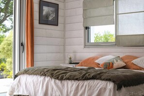 Ein Schlafzimmer mit Bett, einem Fenster mit Vorhängen, einem Nachttisch mit Lampe und einem gerahmten Bild an der Wand.