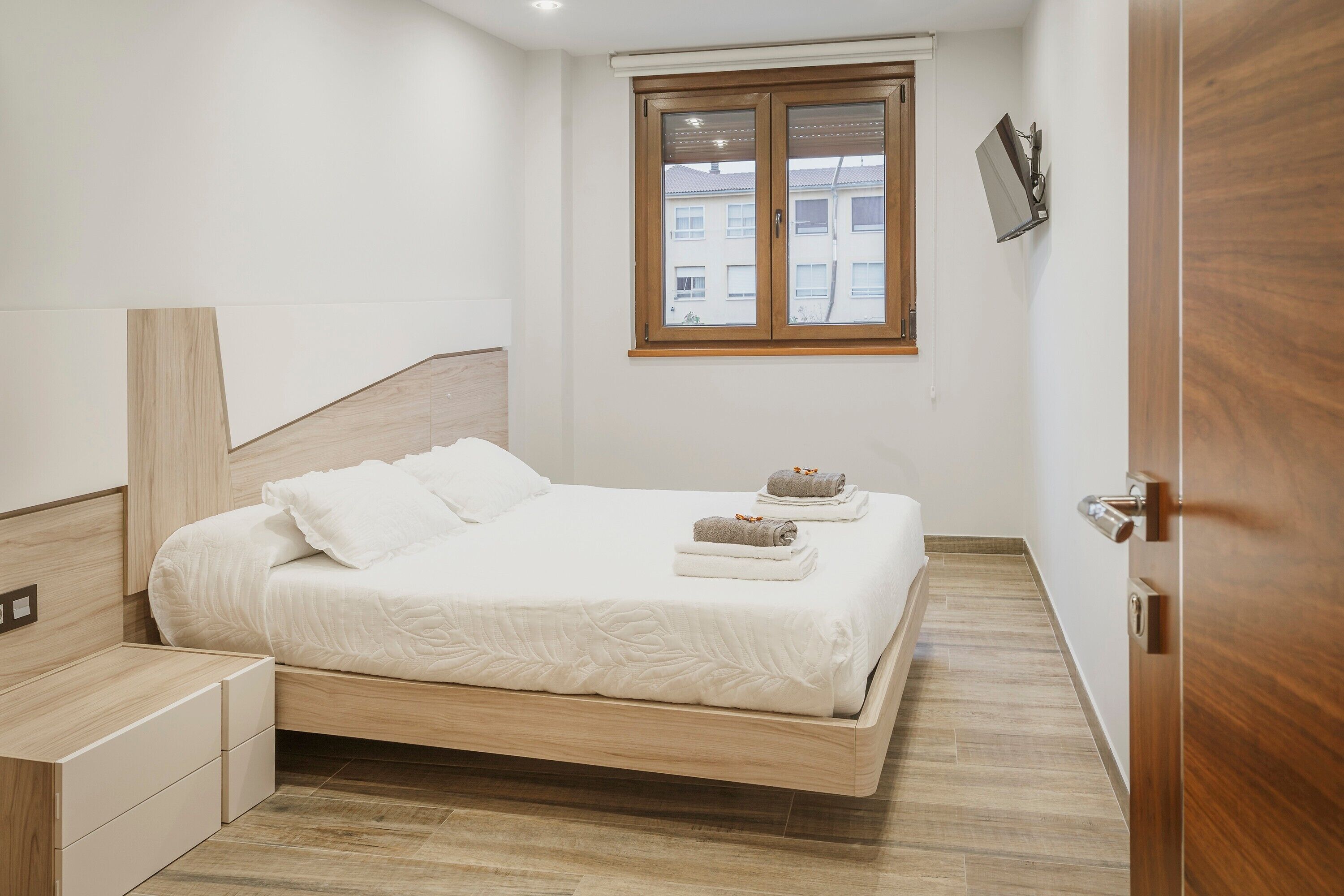 A modern bedroom with a wooden bed, a small bedside table, a nightstand, and a window with a view of buildings.