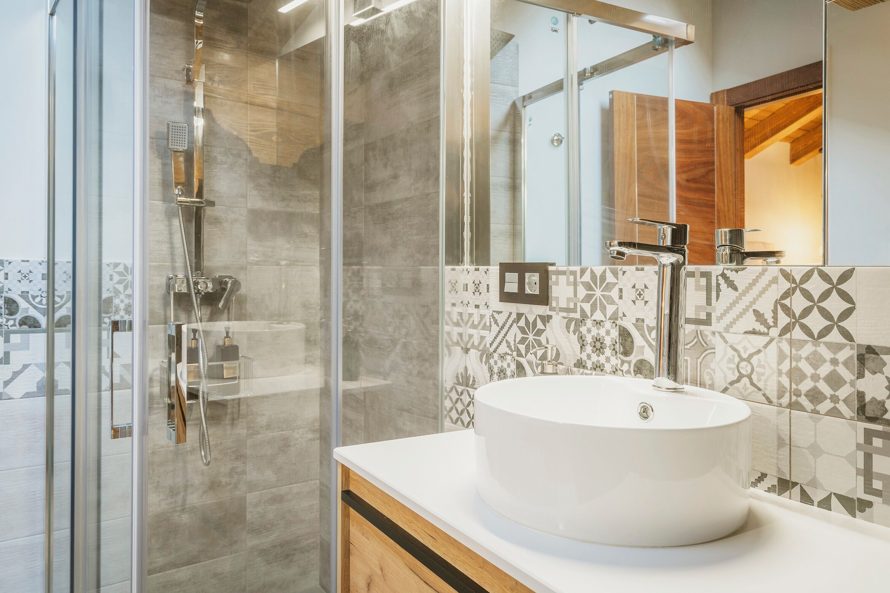 A modern bathroom with a glass-enclosed shower, a round white sink, and patterned tiles.