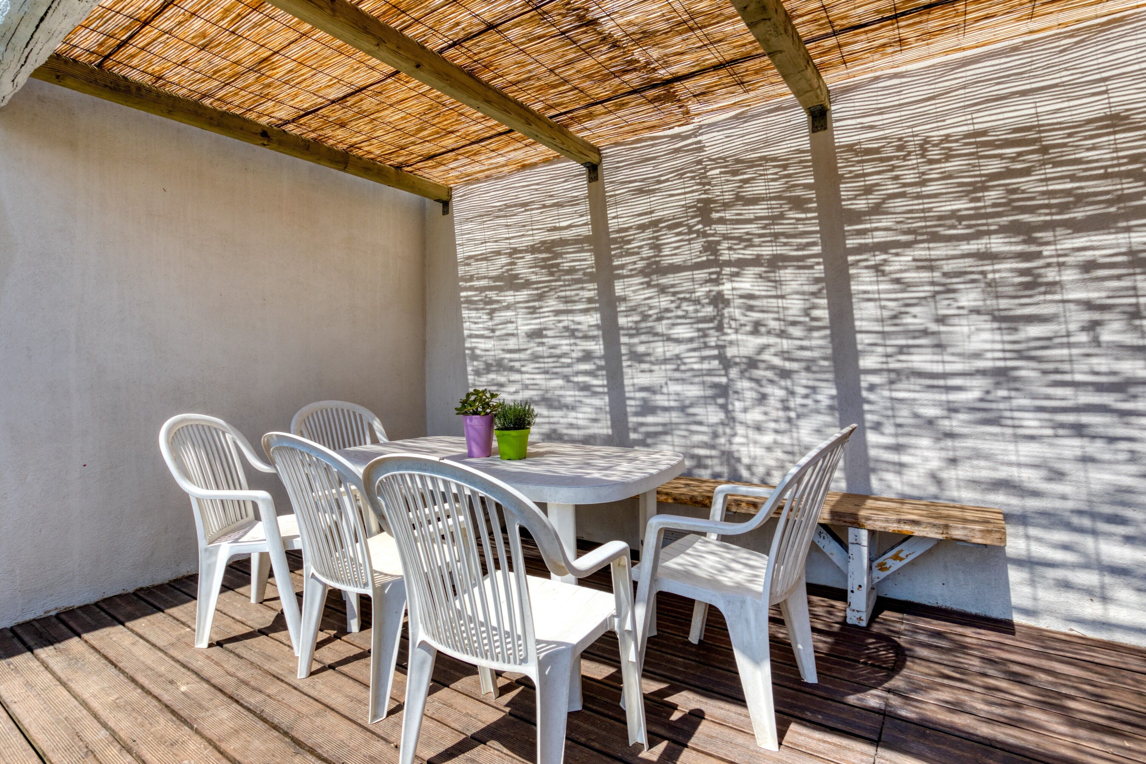 A covered patio with a round table and white plastic chairs, a small potted plant, and a wooden bench.
