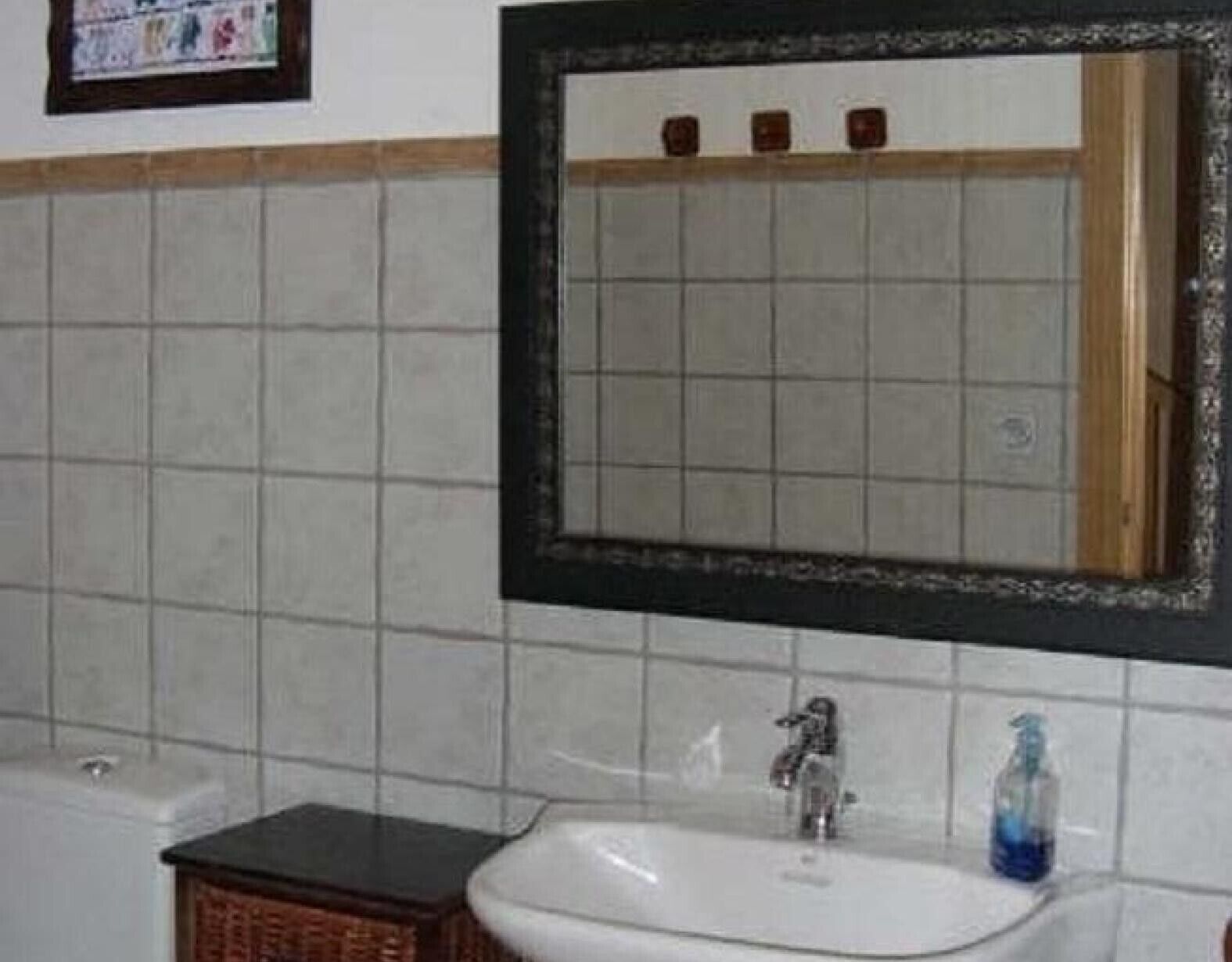 A bathroom with a tiled wall, a white sink, a mirror with a wooden frame, and a small wicker basket.