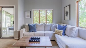 A living room with a light-colored sofa, a wooden coffee table, and large windows offering a view of greenery.