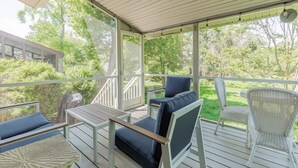 A covered outdoor deck with seating and a table, surrounded by greenery.