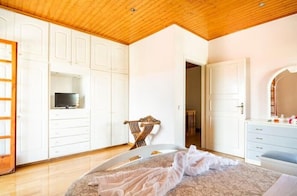 Quarto com teto de madeira, guarda-roupa branco, cama com cobertura branca e televisão em uma prateleira.