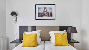 Habitación de hotel con dos camas, cada una con almohadas blancas y amarillas, un cuadro con la Torre Eiffel en la pared y una lámpara en la mesita de noche.