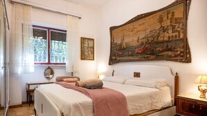 A bedroom with a large bed, a desk, a chair, a window with curtains, and a large painting on the wall.