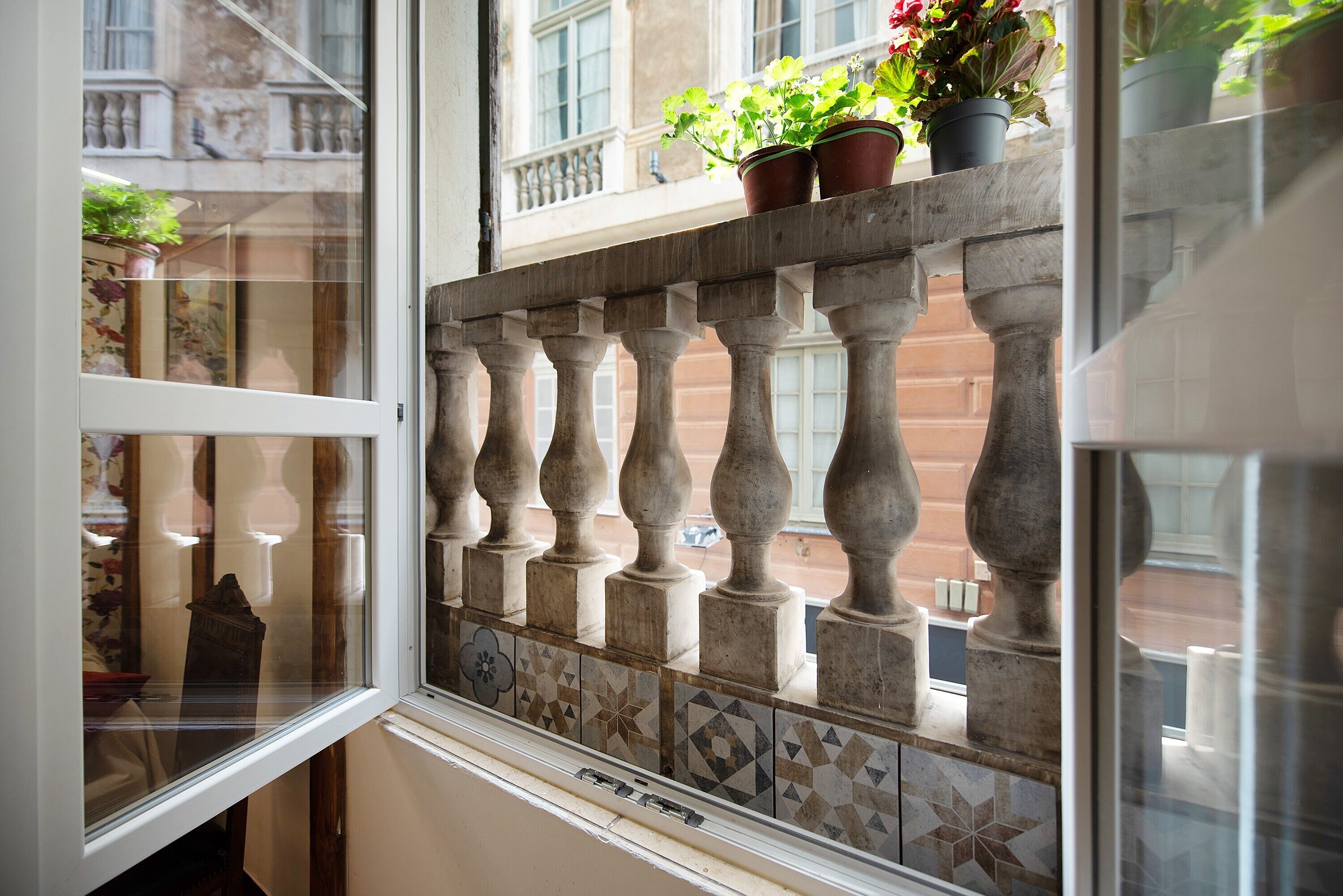 A window with a view of a building's facade, a balcony railing, and potted plants.