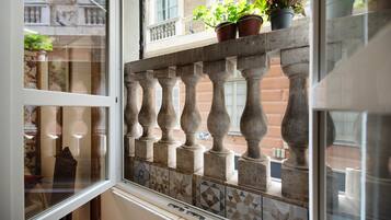 A window with a view of a building's facade, a balcony railing, and potted plants.