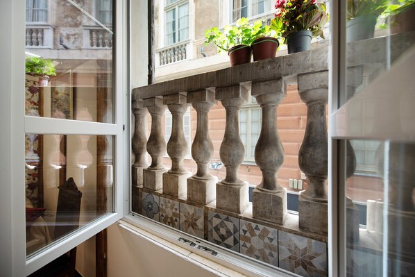 A window with a view of a building's facade, a balcony railing, and potted plants.