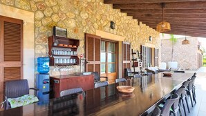 Un área de comedor al aire libre con paredes de piedra, una mesa larga, sillas y un dispensador de agua.