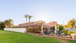 A restaurant with outdoor seating, a white wall, and palm trees. - Apartment 'Las Buganvillas' with Shared Pool, Private Terrace and Air Conditioning (Roquetas de Mar)