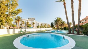 A swimming pool with a surrounding green deck, palm trees, and a building in the background. - Apartment 'Las Buganvillas' with Shared Pool, Private Terrace and Air Conditioning (Roquetas de Mar)