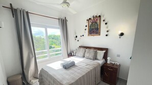 A bedroom with a bed, curtains, a ceiling fan, a nightstand, a wall-mounted lamp, and a window with a view. - 1 Bedroom Apartment in Zanzibar (Zanzibar)