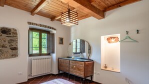 Ein Zimmer mit Holzboden, einer Steinwand, einem Heizkörper, einem Schminktisch, einem Spiegel, einem Fenster und einer Pendelleuchte.