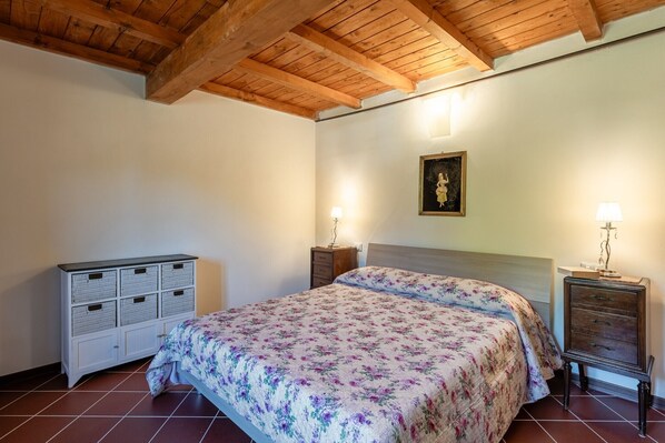 Un dormitorio con techo de madera, una cama con ropa de cama floral, un aparador y una mesita de noche con una lámpara.