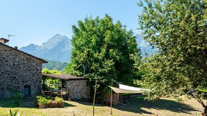 Ein Steinhaus mit überdachter Außenbestuhlung, umgeben von Grünflächen und Bergen im Hintergrund.