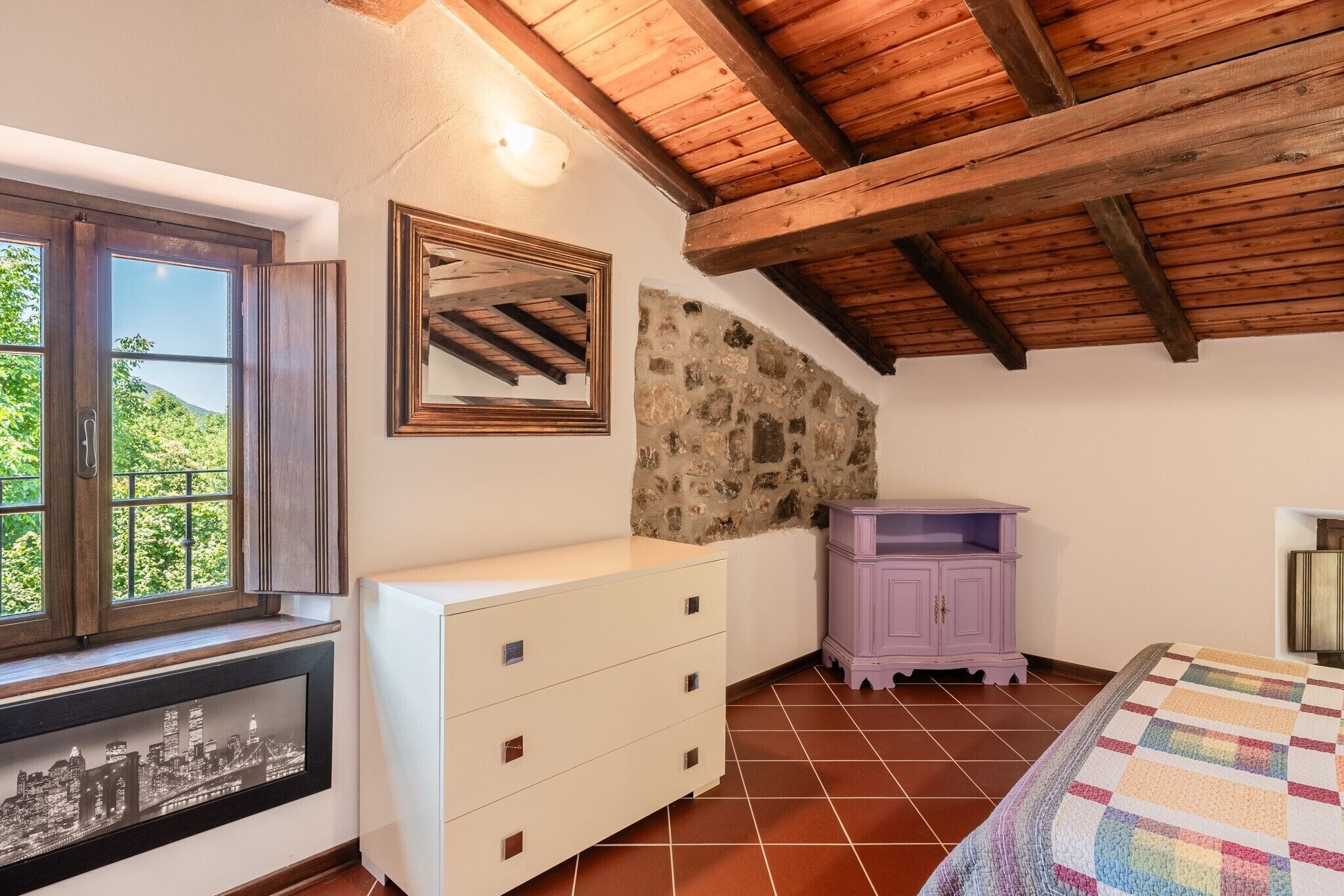 Ein Schlafzimmer mit Stadtbild-Kunstwerk, einer Steinwand, Holzbalken, einem Fenster, einer weißen Kommode, einem violetten Schrank und einer farbenfrohen Steudecke.
