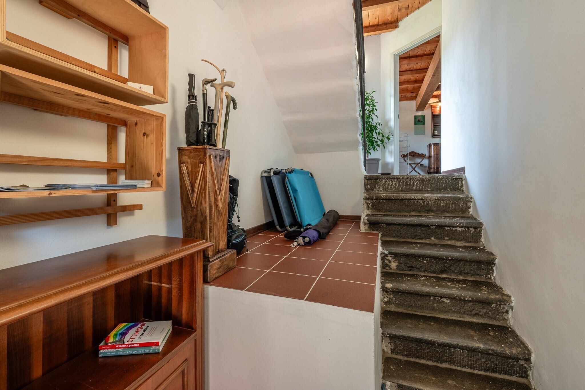 Un escalier menant à l’étage supérieur, des étagères en bois remplies de livres et une valise bleue posée sur le sol.
