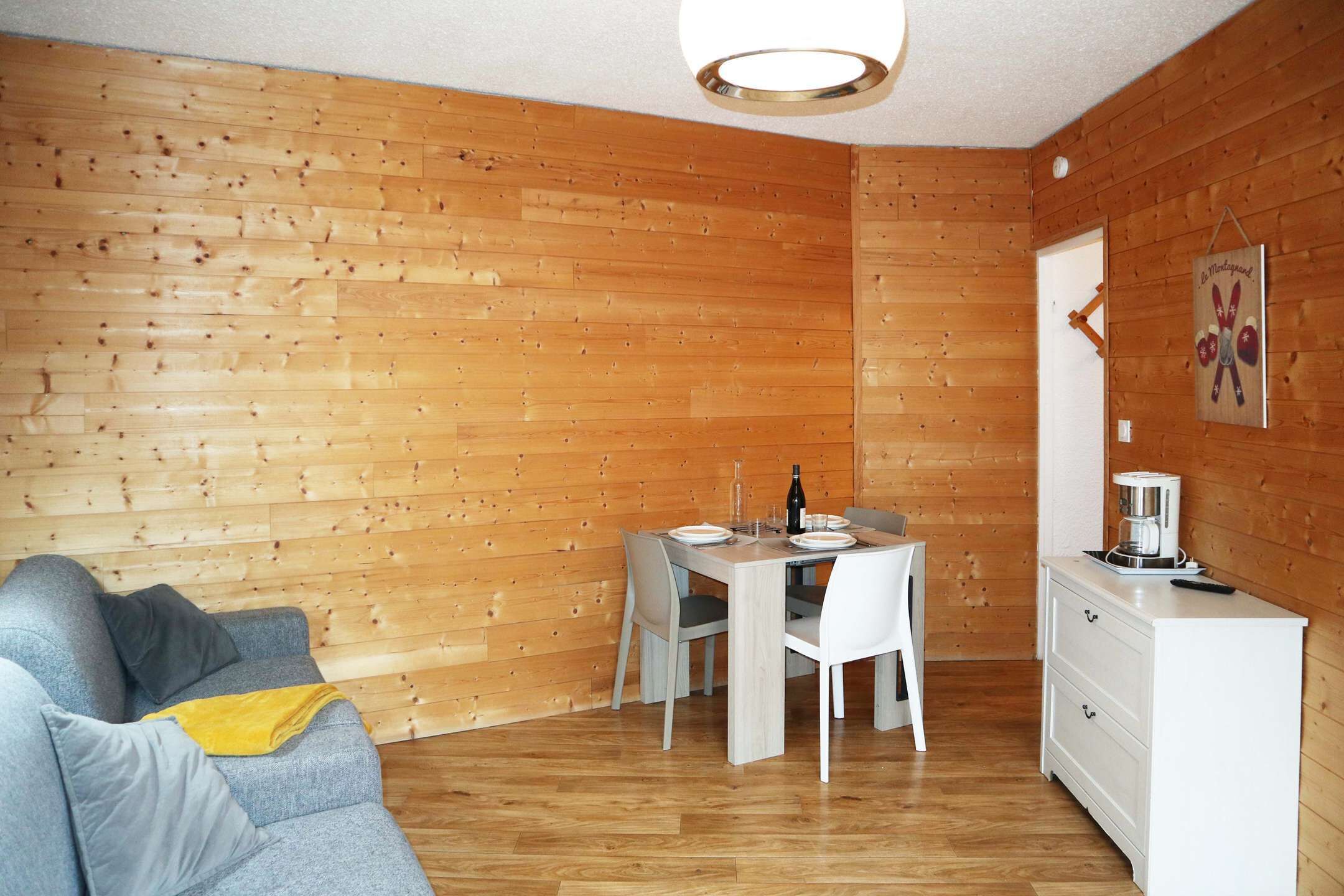 A cozy room with a wooden wall, a dining table with chairs, a white dresser, and a gray sofa with cushions.