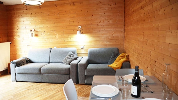 Una stanza accogliente con parete in legno, un divano grigio, un tavolo da pranzo apparecchiato con piatti, bicchieri e una bottiglia di vino.