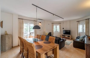 A spacious living area with a wooden dining table, comfortable seating, a flat-screen TV, and large windows.
